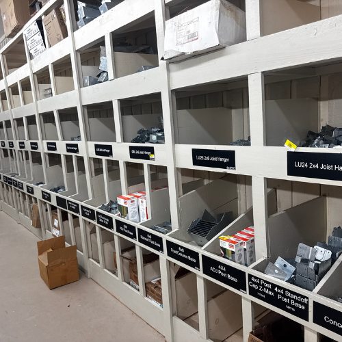 Shelves stocked with various metal hardware and construction items, each labeled with black and white signs in a neatly organized warehouse aisle.
