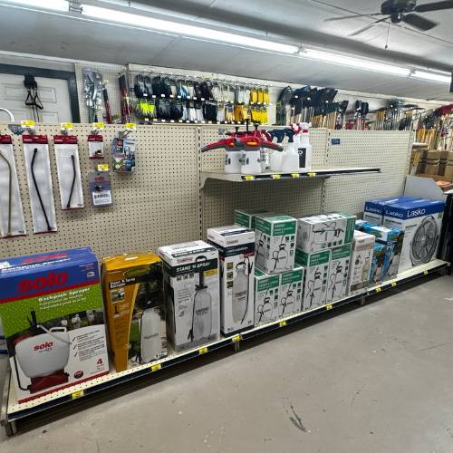 A hardware store aisle with shelves displaying various garden tools and equipment. Items include pruning shears, sprayers, and fans. Bright, organized setting.