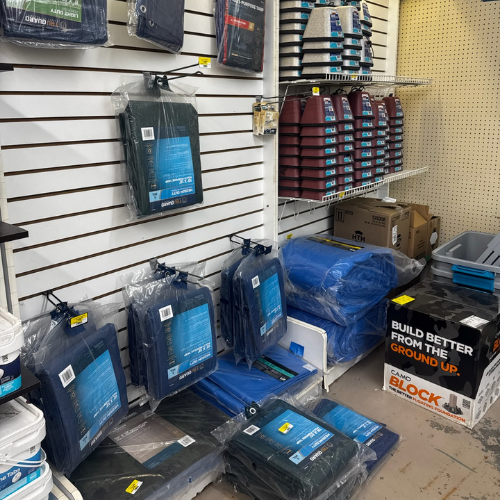 Shelves in a hardware store display stacked tarps, sandpaper packs, and liquid containers. A box labeled 