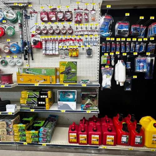 Hardware store aisle displaying various tools and accessories. Shelves are stocked with tapes, fuel cans, garden tools, and other utility items.