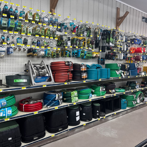 A hardware store aisle displays garden hoses in various colors, hose reels, and tools hanging on a pegboard. The scene is organized and vibrant.