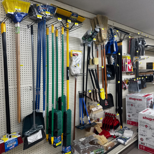 Hardware store aisle with cleaning tools: brooms, mops, dustpans, and brushes hanging on a pegboard. Boxes of Shop-Vac products are on the shelf below.