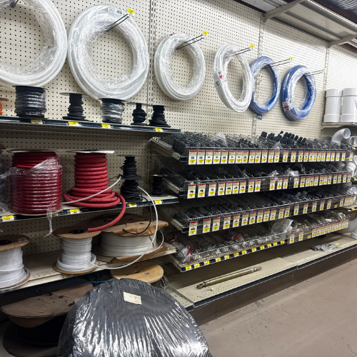 A hardware store aisle displays coiled cables and wires, organized on shelves. Spools of red, black, and white cables are neatly stacked below.