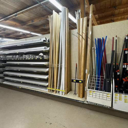 A hardware store aisle with metal and wood strips and rods vertically organized on shelves. Labels indicate 'Ready Cut Pipe'. Bright, organized setting.