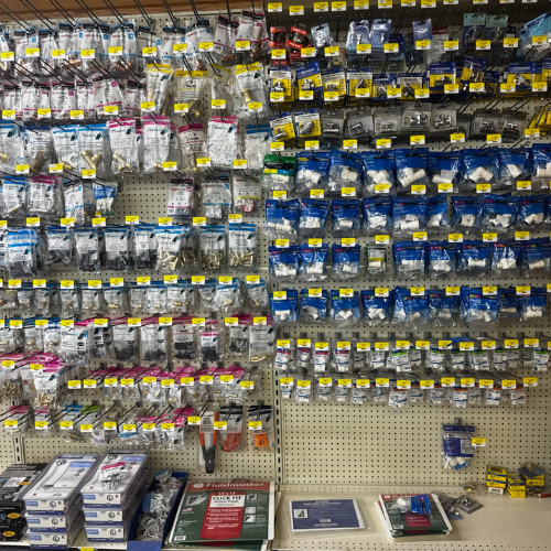 Wall display of hardware items, organized in plastic packages on hooks. Shelves at the bottom hold more items. Neat and colorful with yellow price tags.