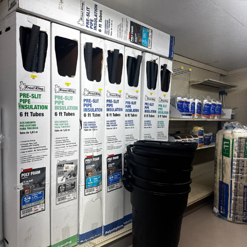 A hardware store aisle displays Frost King pre-slit pipe insulation in tall white boxes. Nearby are stacked black bins and shelves with various supplies. The area is well-lit and orderly, creating a practical, utilitarian atmosphere.