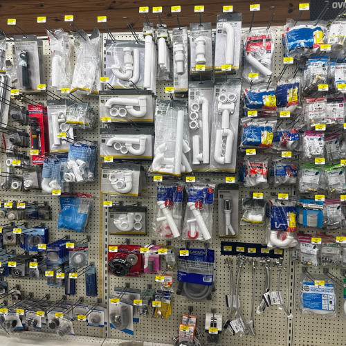 A store wall displays various plumbing supplies in neat rows, including pipes, fittings, and hoses, all in clear packaging with yellow price tags.