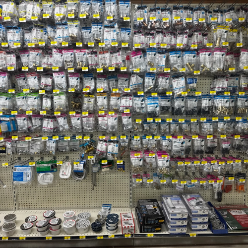 A store aisle displays various packaged hardware items neatly hung on a wall. The shelves below hold small containers and boxes, creating an organized, busy atmosphere.