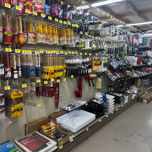 Shelves in a hardware store display a variety of painting tools, including brushes, rollers, trays, and accessories. The setting is organized and well-stocked, creating a practical and utilitarian atmosphere.