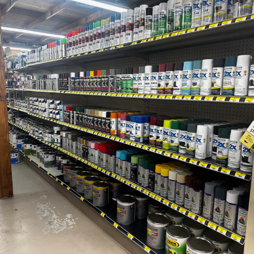 A hardware store aisle with shelves full of various spray paint cans in a spectrum of colors and metal paint cans below, creating a vibrant, organized display.