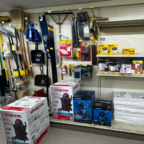 Hardware store aisle displaying various cleaning and tool supplies, including brooms, rakes, Shop-Vac boxes, and packaged filters on pegboard shelving.