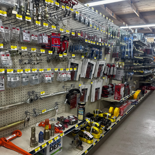 A hardware store aisle with pegboard displays various tools and hardware items, including hooks, pipes, and straps. Bright fluorescent lighting.