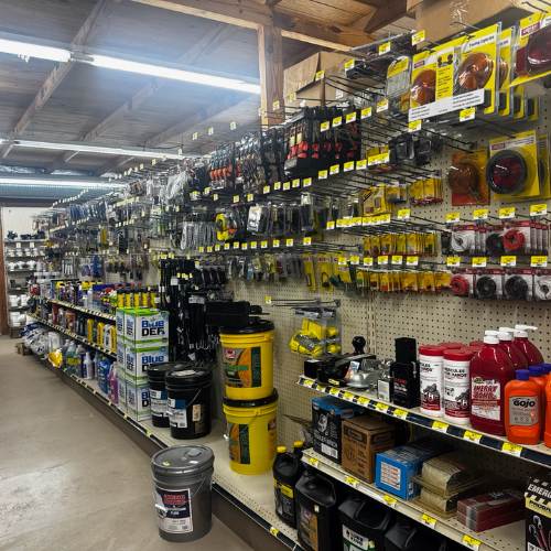 A hardware store aisle displays neatly organized tools, cleaning supplies, and buckets on shelves. Bright lighting creates an orderly and welcoming atmosphere.