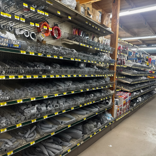 A hardware store aisle with shelves full of metal pipes, fittings, and tools. The scene is neatly organized and brightly lit, conveying a sense of order.
