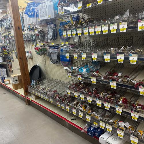 A hardware store aisle with shelves of padlocks, packaged cables, and chains. Labels display prices. The floor is concrete, creating an organized, utilitarian feel.