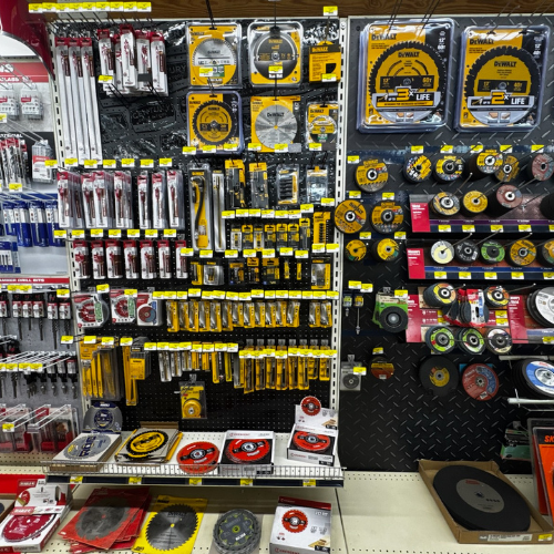 A hardware store aisle featuring a wall filled with various saw blades, drill bits, and tools. Bright packaging, predominantly yellow and black, signifies different brands. The display is neatly organized, conveying a sense of variety and readiness for DIY projects.