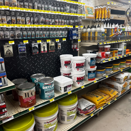 Shelves in a hardware store display various construction supplies, including sealants, paints, adhesives, and bags of concrete mix. Bright lighting highlights the organized assortment.