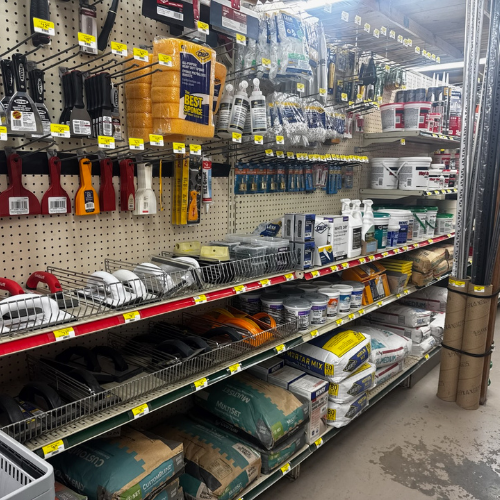 Hardware store aisle with shelves of tools, paint supplies, and construction materials. Items are neatly organized. Bright lighting, busy atmosphere.