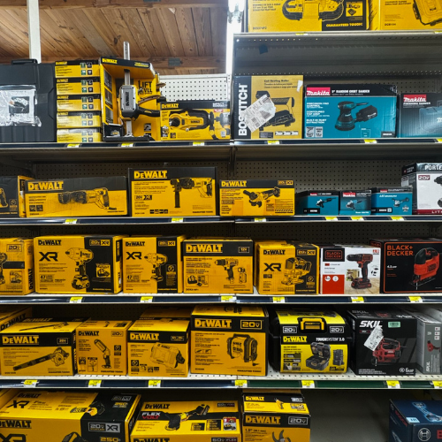 A store shelf displaying a variety of power tools in bright yellow DeWalt boxes, with some Makita and Black & Decker items, creating a hardware-themed atmosphere.