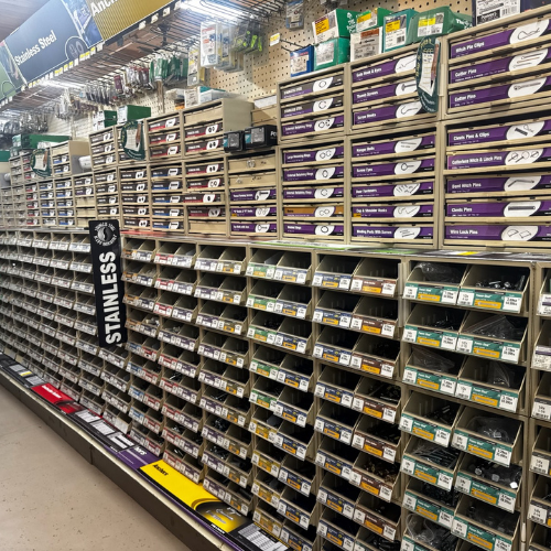 A hardware store aisle with neatly organized rows of metal fasteners, screws, and small parts in labeled boxes. Overhead signs indicate 