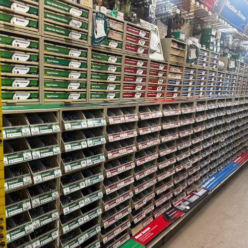 A wall of organized hardware store shelves filled with boxes of various screws. Labels in green, red, and blue. Neat and orderly display.
